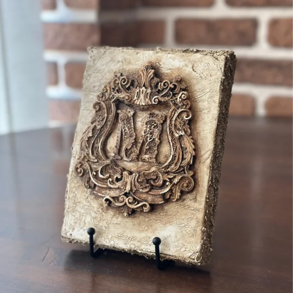 Vista lateral del escudo con inicial M en relieve, acabado hueso antiguo, serie Memor, colección Emblemata.